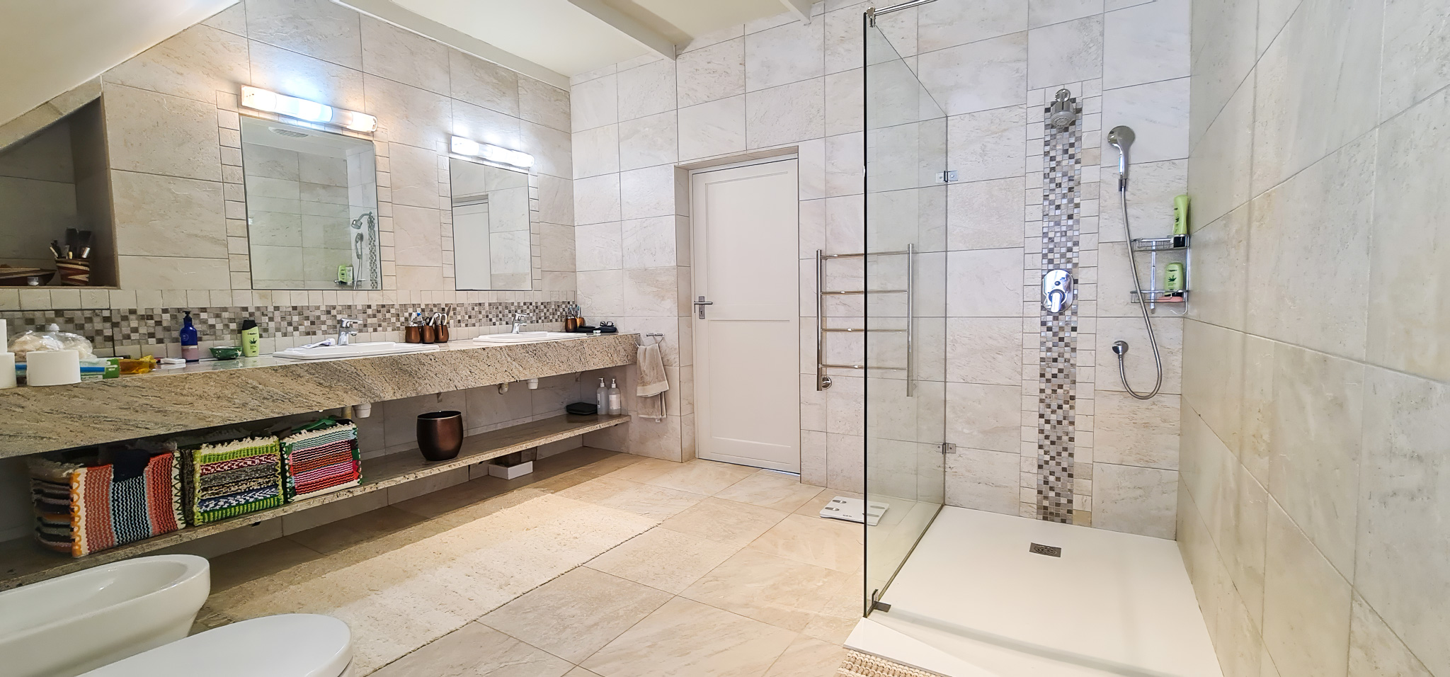 Interior Spaces Elegant bathroom featuring natural stone tiles, glass shower enclosure with mosaic tile accents, granite vanity with modern fixtures, and sophisticated lighting