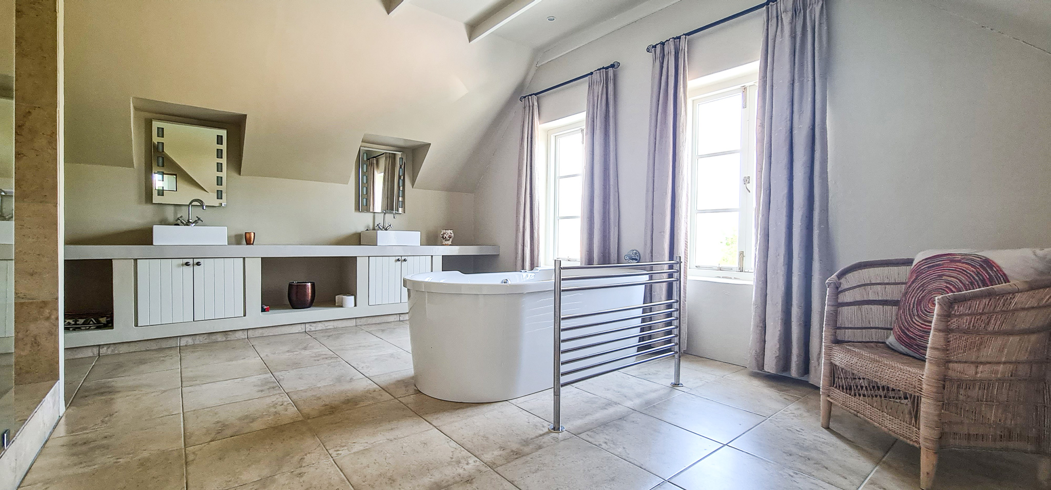 Interior Spaces Luxurious bathroom featuring exposed beam ceiling, freestanding bathtub, dual vessel sinks on custom cabinetry, natural stone flooring, and large windows with natural light