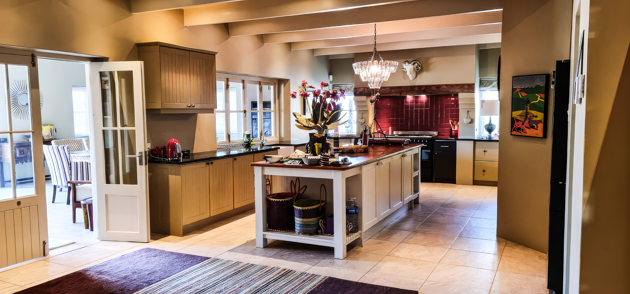 Interior Spaces Modern kitchen interior with wood cabinetry, exposed beams, crystal chandelier, and red tile backsplash