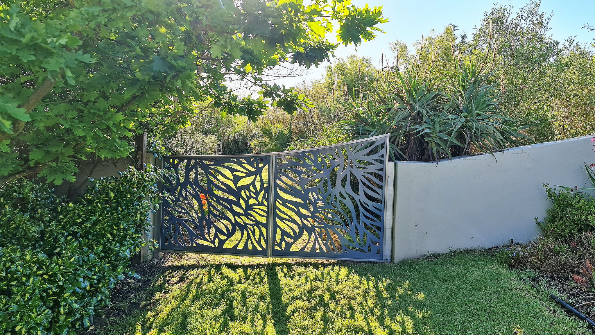 Gardens & Grounds Decorative metal gate with intricate leaf pattern design surrounded by lush green landscaping and mature gardens