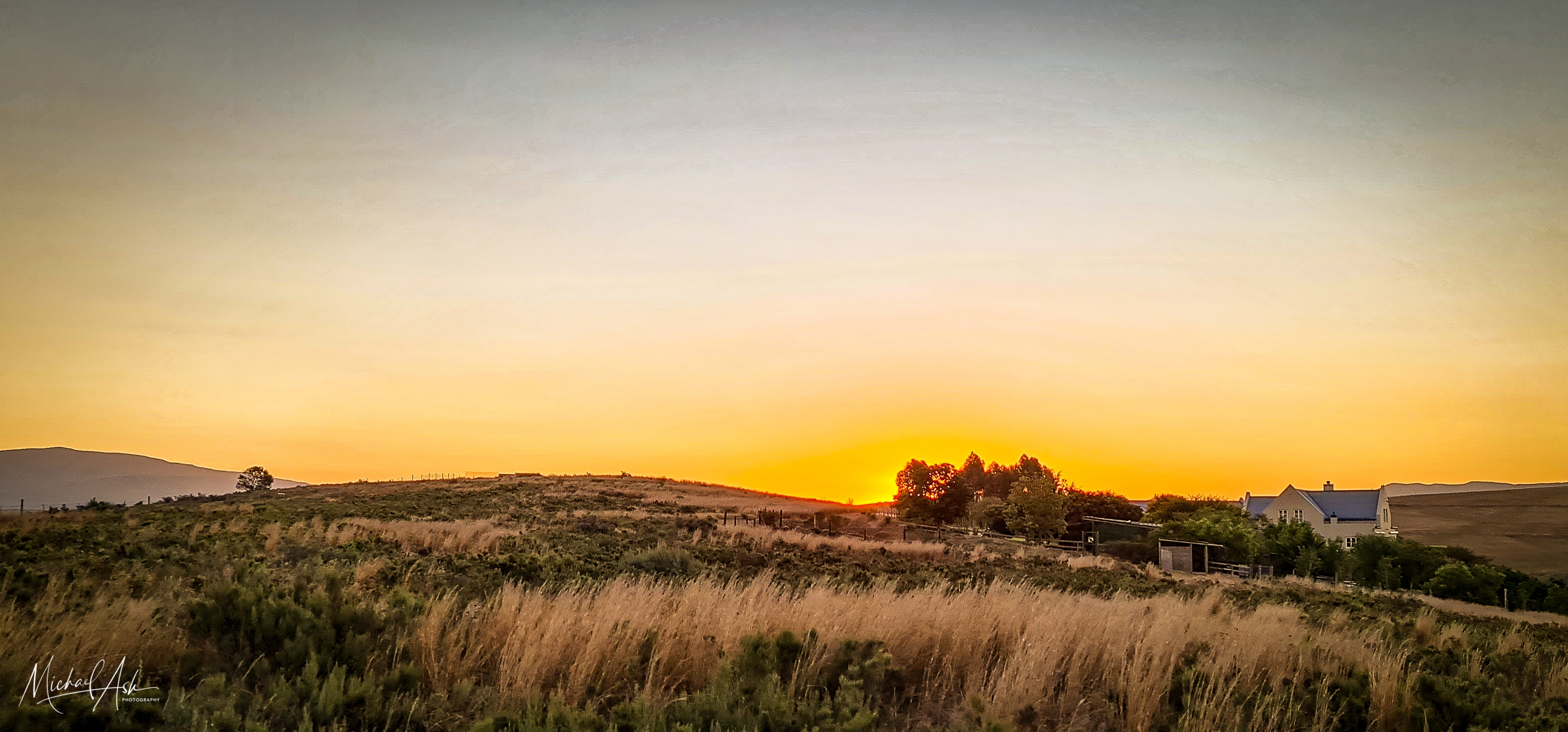 Landscape & Views Golden hour landscape showing native Renosterveld vegetation, rolling countryside, and property buildings in the distance under dramatic sunset lighting