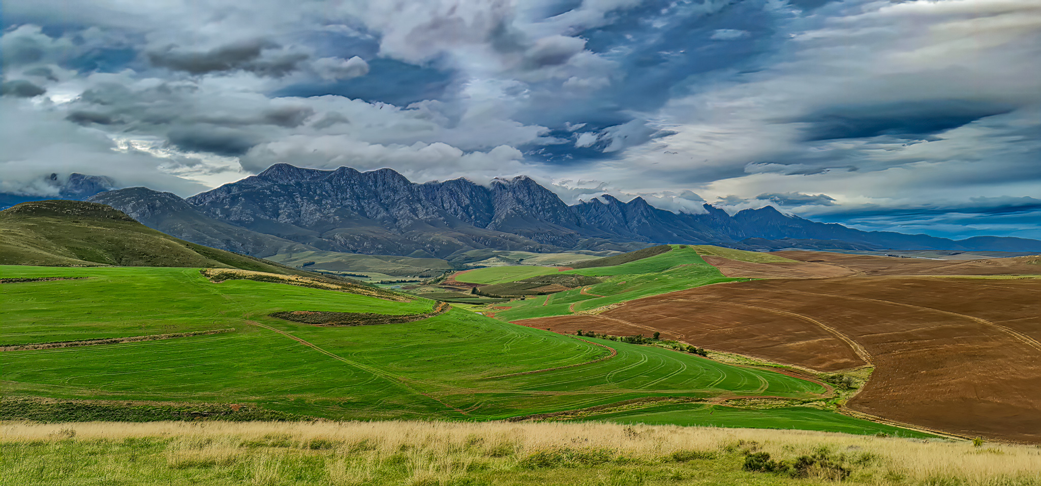 Landscape & Views Winter landscape showing dramatic cloudy sky, layered mountain ranges, and lush green agricultural fields with native vegetation